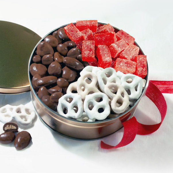 Assorted candies including chocolate covered almonds, red jelly cubes, and white pretzels in a metal tin on a white background.